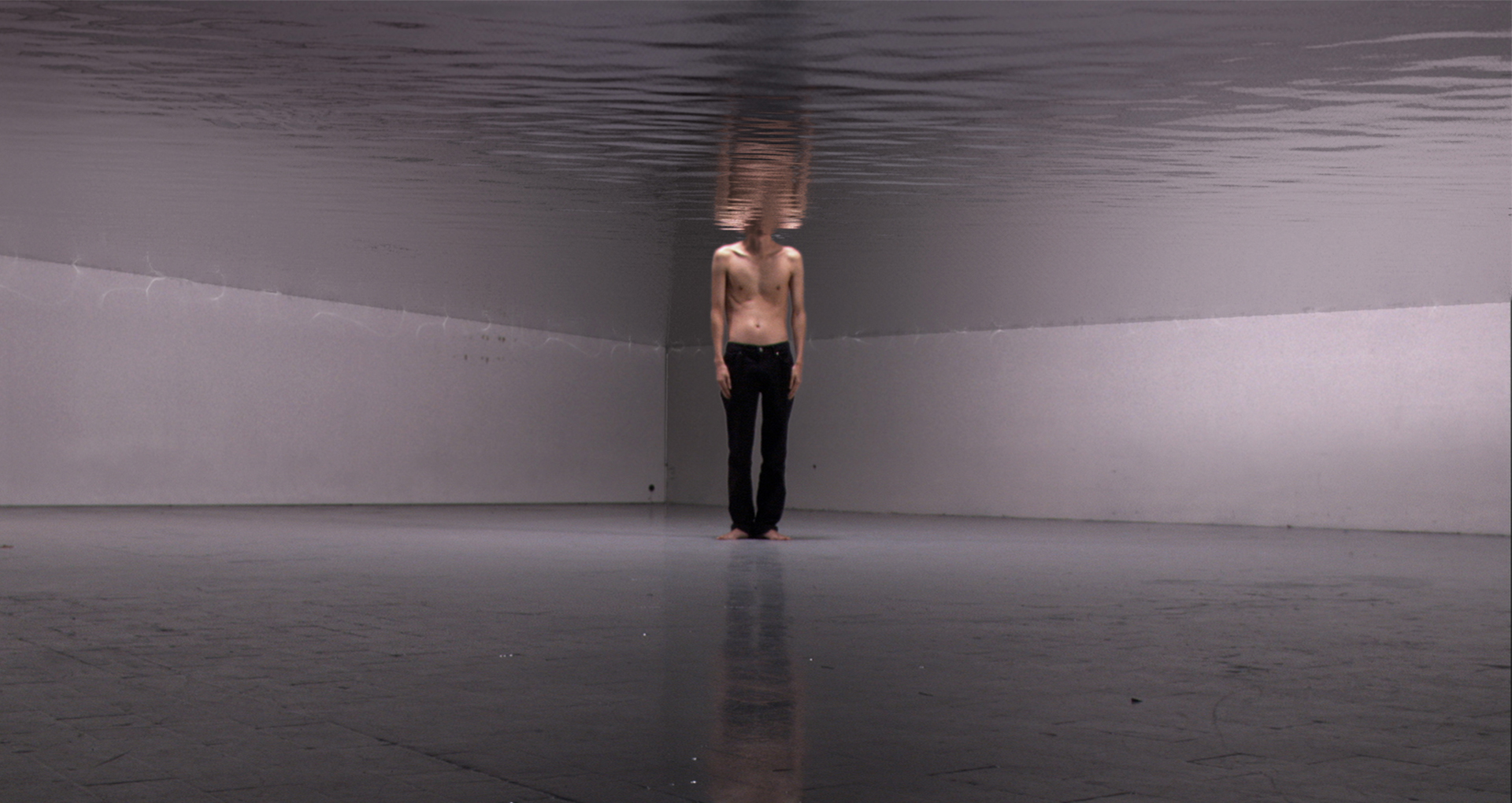 Montage vidéo. Un homme debout torse-nu avec un pantalon noir au milieu d’une pièce. La moitié supérieure de la pièce est immergée dans l’eau et la tête du personnage aussi.