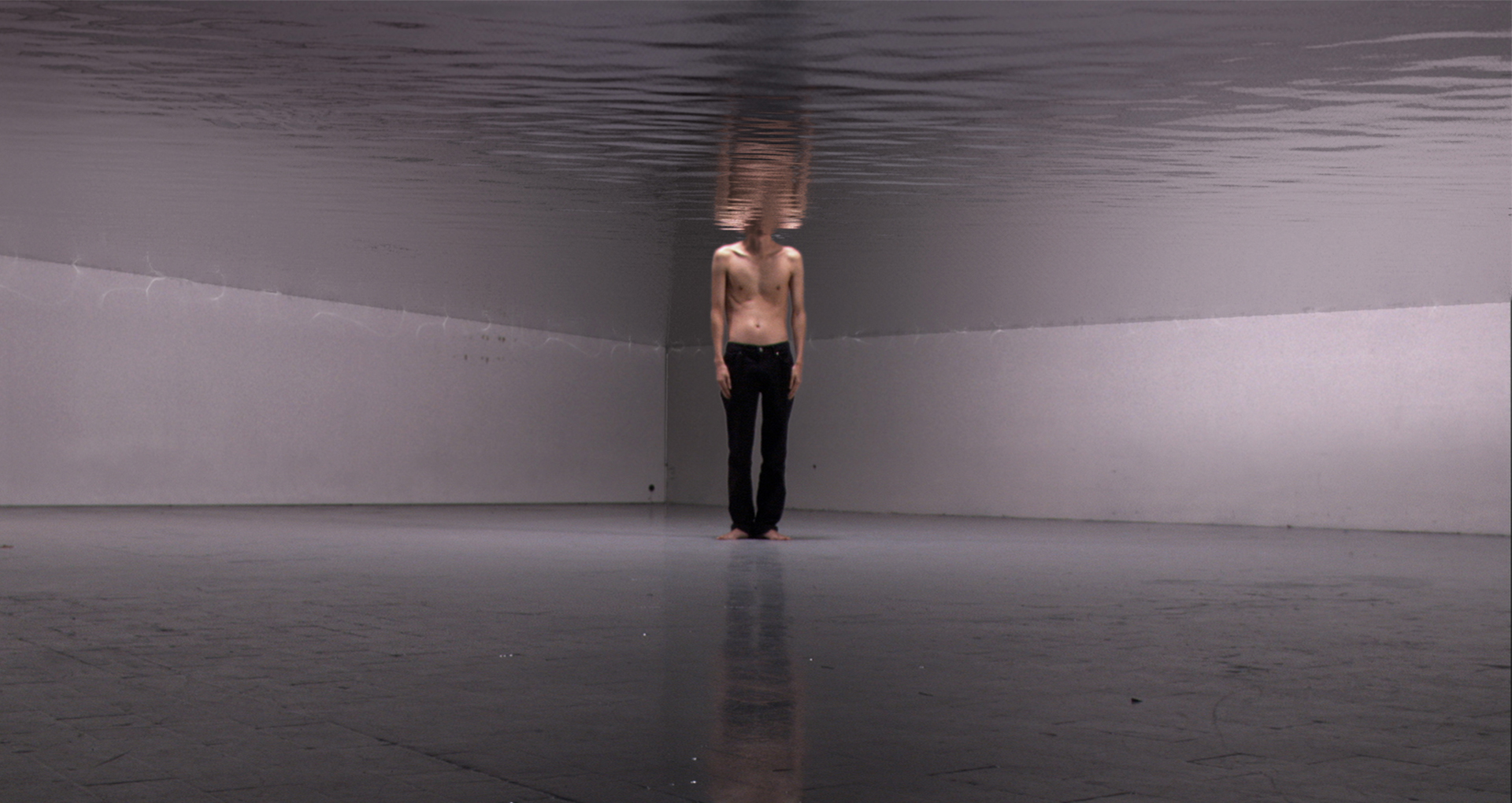 Grecu / Mihal Coagulate Montage vidéo. Un homme debout torse-nu avec un pantalon noir au milieu d’une pièce. La moitié supérieure de la pièce est immergée dans l’eau et la tête du personnage aussi.